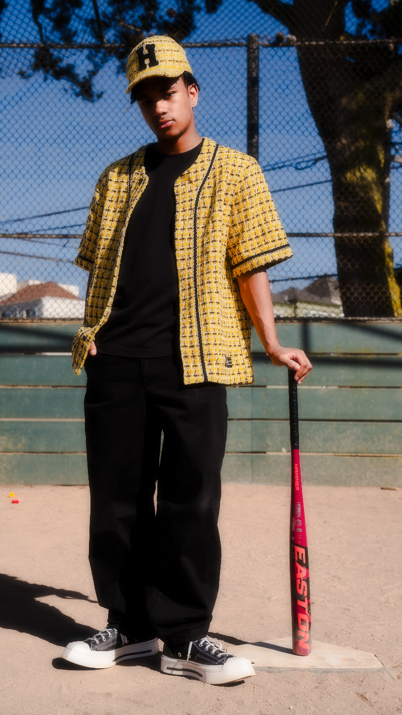 Person wearing a yellow checkered shirt and black pants, holding a red baseball bat on a sports field.