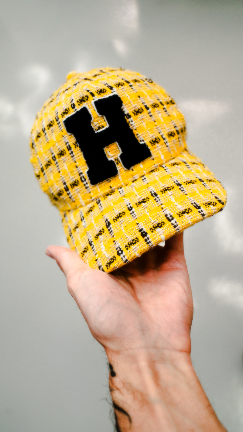 Yellow checkered cap with a black letter 'H' held by a hand against a gray background

